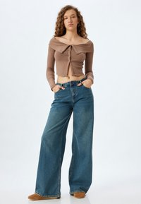 Brun ribbet cropped skjorte med off-shoulder design og knapper, parret med blå vidde jeans og beige fodtøj. Lys baggrund.