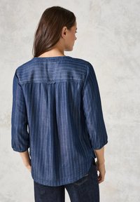 Camicia in denim scuro con strisce bianche verticali, orlo curvo e maniche a tre quarti. Presenta una piega e dettagli di cucitura sottili.