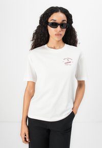 Donna con lunghi capelli ricci, che indossa occhiali da sole neri, una t-shirt bianca del Billabong Surf Club e pantaloni neri, in piedi contro uno sfondo bianco.