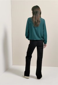 Blusa teal a maniche lunghe realizzata in tessuto morbido, abbinata a pantaloni neri a zampa d'elefante. Si possono notare un design retro discreto e una vestibilità rilassata.