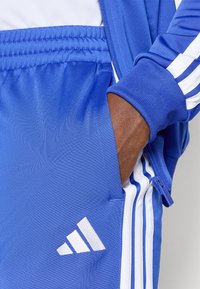 Modrá športová bunda s bielymi pruhmi a zipsovými vreckami, ktorá má jemnú textúru a výrazné biele logo na hrudi.