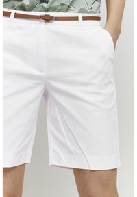 Bílé šortky ke kolenům se štíhlým hnědým páskem, nošené s jednou rukou zastrčenou v pravé kapse.