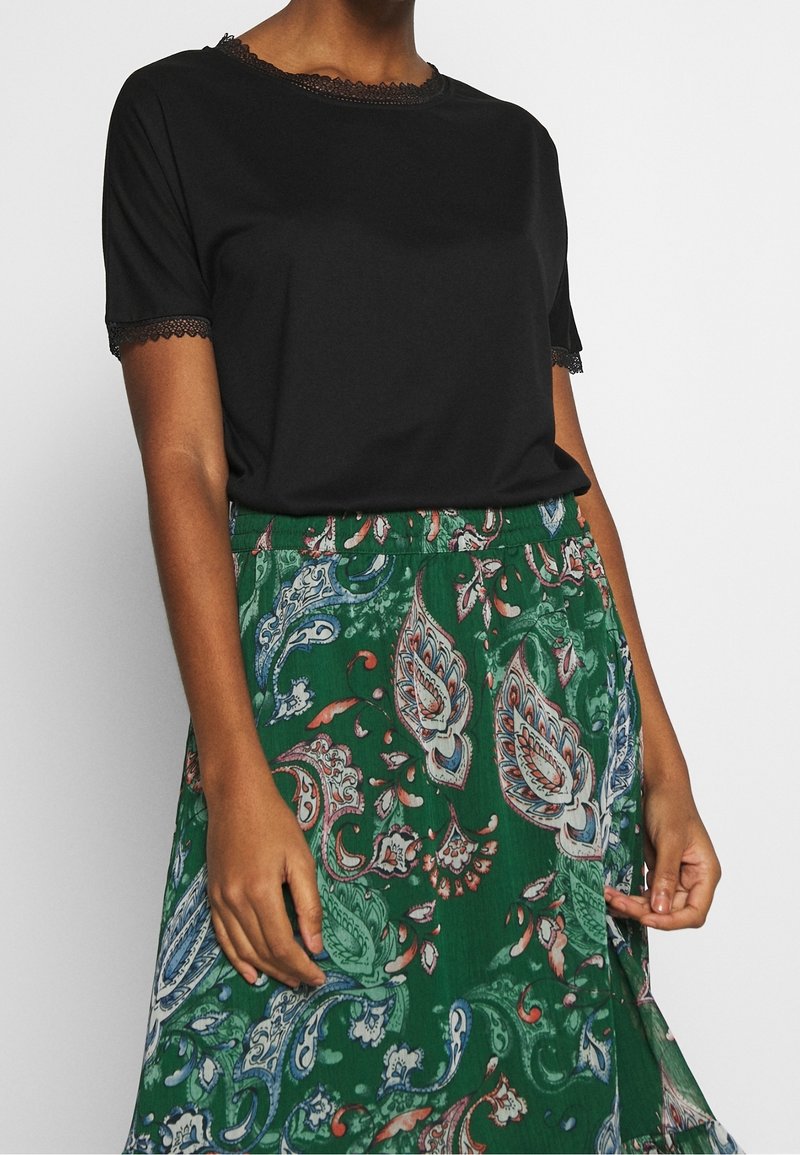 Femme portant un haut noir à manches courtes avec bordure en dentelle et une jupe verte à motif cachemire.