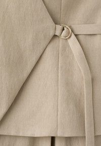 Tissu beige plié avec une ceinture sécurisée par une boucle double anneau argentée, montrant une texture et des coutures détaillées.