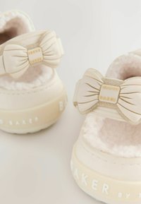 Chaussures beige clair avec un intérieur doux et moelleux, ornées d'un accent de nœud proéminent et du nom de marque gravé "Baker" sur la semelle.