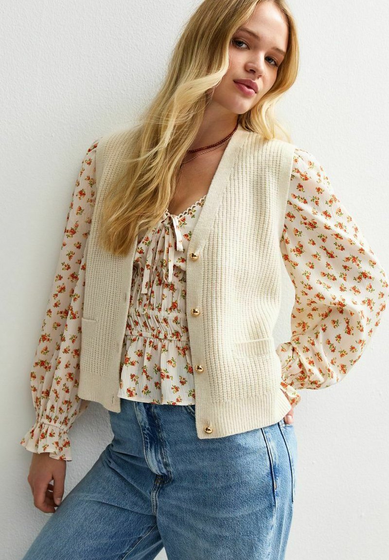 Junge Frau mit langen blonden Haaren, die einen cremefarbenen Strickpulli über einer blumigen Bluse und blauen Jeans trägt, steht vor einer weißen Wand.