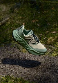 Green and cream running shoe featuring a textured upper, black laces, prominent 'N' logo, and a cushioned sole, suspended above a forest floor.