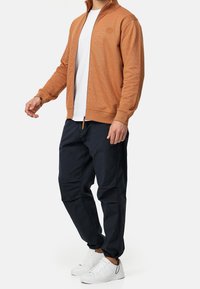 Veste à capuche zippée en tissu texturé orange, associée à un pantalon cargo bleu marine foncé et des baskets blanches ; arbore un logo sur la poitrine.