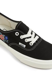 Fekete vászon sneaker fehér gumi talppal és fűzővel. Virágmintás hímzés található az oldalán, valamint egy steppelt minta a szélein.