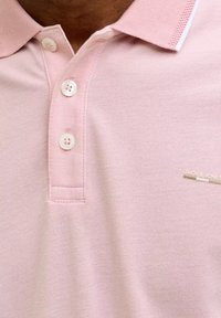 Nahaufnahme einer Person, die ein hellrosa Poloshirt mit drei weißen Knöpfen und einem kleinen "Jack & Jones"-Logo auf der Brust trägt.