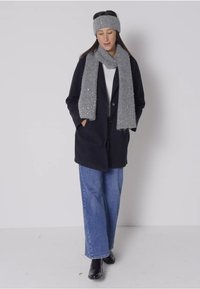 Cappotto nero, sciarpa grigia lavorata a maglia con paillettes, top bianco, jeans larghi blu, stivaletti neri, indossato con le mani nelle tasche.