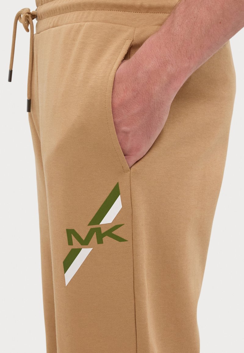Pantalones de chándal beige con un bolsillo lateral, con un diseño gráfico en verde y blanco y las iniciales "MK" en la pantorrilla. Textura suave.