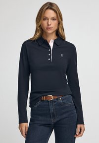 Langärmliges dunkles Poloshirt mit pinkfarbenen Kragenakzenten, vier Knöpfen an der Blende und kleinem Logo. Kombiniert mit hochtaillierten dunklen Jeans und Gürtel.