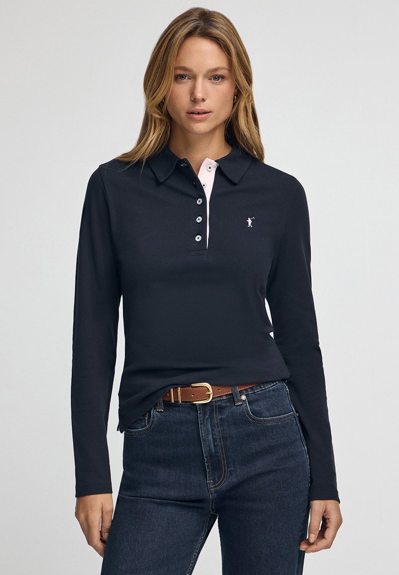 Langärmliges dunkles Poloshirt mit pinkfarbenen Kragenakzenten, vier Knöpfen an der Blende und kleinem Logo. Kombiniert mit hochtaillierten dunklen Jeans und Gürtel.