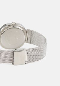 Sidabrinė tinklinė laikrodis su apvaliu korpusu ir kontrastinga lygiąja nugarėle. Yra metalinė sagtis su prekės ženklo ženklu, demonstruojanti elegantišką, minimalistinį dizainą.