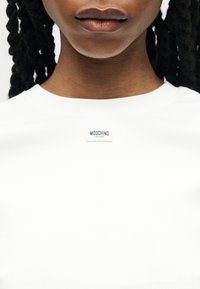 Κοντινό πλάνο προσώπου με σκούρα πλεγμένα μαλλιά που φοράει λευκή μπλούζα με μικρή ετικέτα Moschino Jeans στον λαιμό.