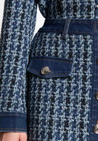 Vestido de denim y tejido texturizado con un patrón azul y negro, que presenta detalles en botones metálicos y una cintura ceñida con cinturón.