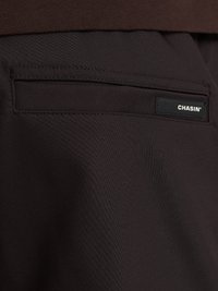 Dettagli in tessuto nero dei pantaloni con una tasca posteriore e un'etichetta rettangolare con la scritta "CHASIN'" in testo bianco. Texture liscia e opaca.