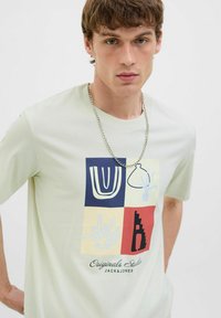 Lys grøn bomulds t-shirt med et flerfarvet geometrisk grafik med abstrakte former og teksten "Originals Studio JACK & JONES" i bunden.
