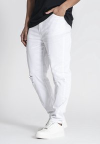 Vaqueros blancos de denim con un ajuste relajado, que presentan detalles desgastados en las rodillas y los bolsillos laterales, combinados con zapatillas blancas.