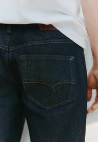 Sötétkék farmernadrág hímzett hátsó zsebbel, kontrasztos világosbarna cérnával és bőrbőrrel a derékrészen. Egyszerű kialakítás.