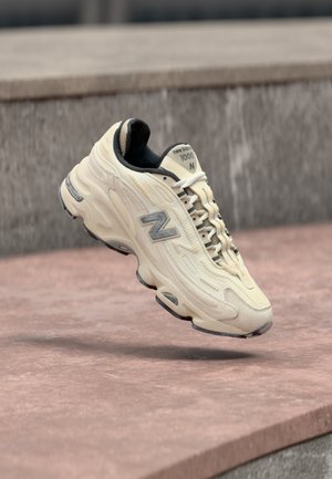 Scarpa da ginnastica New Balance 1000 color crema con lacci bianchi, logo grigio e interno nero, mostrata in aria sopra una superficie di pietra.