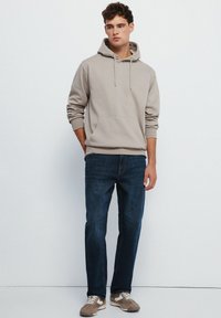 Beige Kapuzenpullover mit einer Fronttasche und Kordelzügen, kombiniert mit dunkelblauen Jeans und braunen Sneakers mit weißen Akzenten.