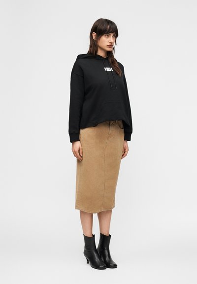 Γυναίκα που φοράει μαύρο φούτερ με κουκούλα, μπεζ midi φούστα και μαύρες μποτάκια μέχρι τον αστράγαλο, στέκεται μπροστά σε απλό φωτεινό φόντο.