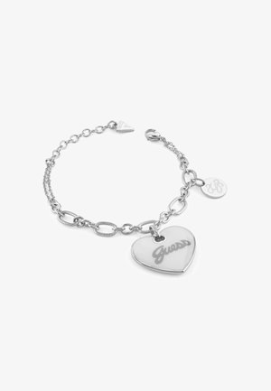 Pulsera de plata con eslabones de cadena interconectados. Presenta un charm en forma de corazón grabado con "Guess" y una pequeña etiqueta redonda.