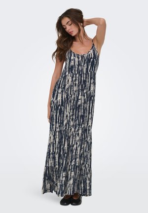 Femme posant dans une robe sans manches, longue jusqu'au sol, à motif tie-dye bleu marine et blanc, avec des ballerines noires sur un fond uni.