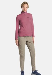 Veste en polaire couleur rose poussiéreux, fermeture éclair intégrale, col montant, texture lisse, deux poches latérales ; associée à un pantalon beige clair et des chaussures de couleur marine.