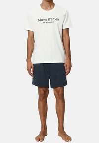 Fehér pamutpóló fekete feliratos logóval az elején, sötétkék rövidnadrággal párosítva. Egyszerű, kényelmes szabás, rövid ujjú, hétköznapi kialakítás.