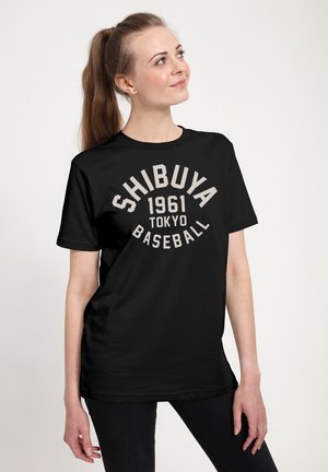 Ung kvinde med lang hestehale iført en sort T-shirt med teksten "Shibuya 1961 Tokyo Baseball," stående foran en hvid baggrund.
