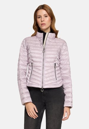 Woman wearing a light purple quilted zip-up jacket with two zippered pockets, paired with black jeans, standing against a plain background.