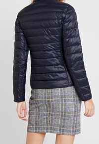 Veste matelassée en duvet bleu marine avec un col haut et des manches longues, présentant une texture lisse et des coutures horizontales, associée à une jupe à carreaux.