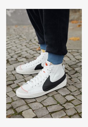 White high-top sneakers with black leather accents, textured upper, and contrasting soles. Features laces and retro logo detail.