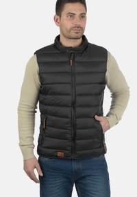 Schwarze, bauchige Weste mit hohem Kragen, Reißverschluss vorne und Lederakzenten. Getragen über einem hellen Pullover und kombiniert mit blauen Jeans.