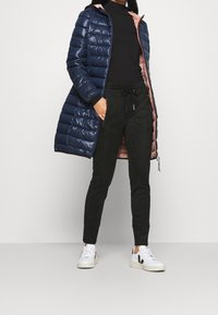 Marinblå puffjacka med en shiny finish och rosa foder, svarta joggingbyxor och vita sneakers med svarta detaljer. Har fickor med dragkedja och huva.