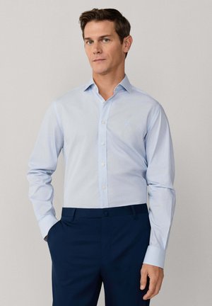 Hombre vistiendo una camisa de vestir de manga larga azul claro metida en pantalones azul oscuro, con una mano en el bolsillo, fondo neutro.