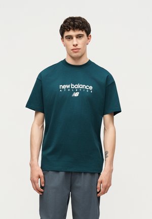Jeune homme aux cheveux courts et bouclés portant un t-shirt bleu sarcelle New Balance Athletics et un short gris, debout devant un fond clair uni.