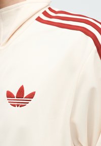 Veste de sport blanche avec le logo Adidas trefoil rouge brodé sur la poitrine et deux bandes rouges le long de la couture de l’épaule.