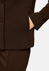 Brauner maßgeschneiderter Anzugjacke mit einer Tasche, die eine glatte Textur und klare Linien aufweist. Knopfverschluss mit detaillierter Stickerei.