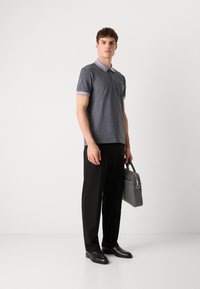 Grå mønstret polo skjorte med lyseblå krage, kombinert med svarte bukser og svarte sko. Modellen holder en grå håndveske.