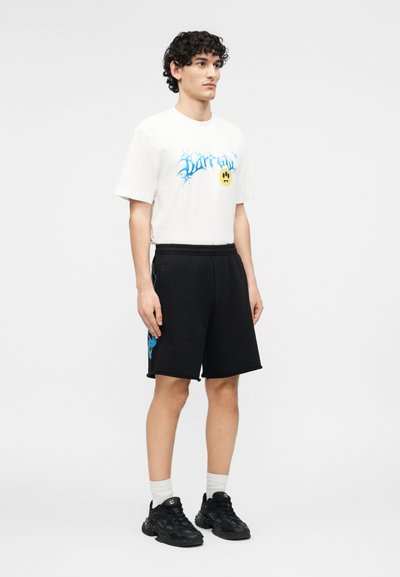 Νέος άνδρας με σγουρά μαλλιά που φοράει λευκό μπλουζάκι με σχέδιο, μαύρα σορτς με μπλε σχέδιο, λευκές κάλτσες και μαύρα αθλητικά, στέκεται μπροστά σε λευκό φόντο.