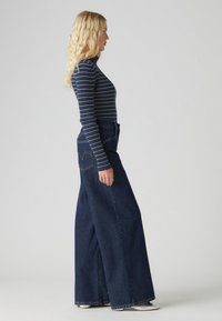 Un haut à manches longues rayé bleu marine et blanc associé à un jean bleu à taille haute et jambes larges, avec un détail de logo sur la poche arrière.