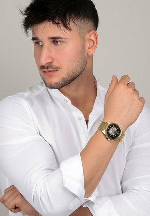 Jeune homme en chemise blanche regardant vers la gauche, portant une montre-bracelet en or avec un cadran noir et doré sur son bras gauche levé.