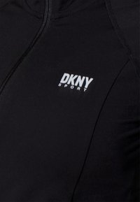 Svart atletisk jacka med dragkedja, DKNY Sport-logotyp i vitt, slät textur och figurnära design.