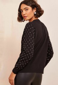 Pulover negru cu un design texturat cu buline pe mâneci, siluetă ajustată, manșete și tiv cu nervuri, purtat peste colanți negri strâmtați.