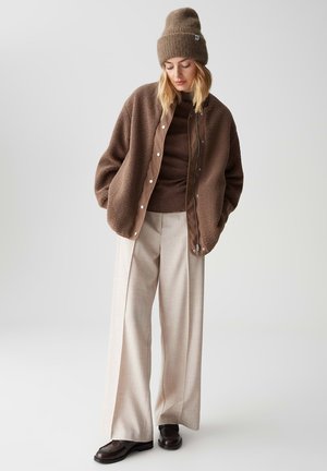 Brown fleece jacket with snap buttons, brown knit beanie, and fitted brown top paired with light plaid wide-leg trousers and brown shoes.