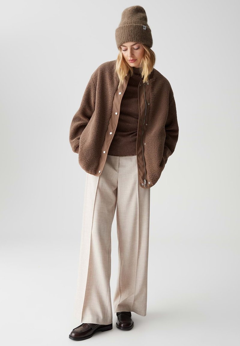 Braune Fleece-Jacke mit Druckknöpfen, braune Strickmütze und enganliegendem braunem Oberteil kombiniert mit hellen karierter weitem Hosen und braunen Schuhen.